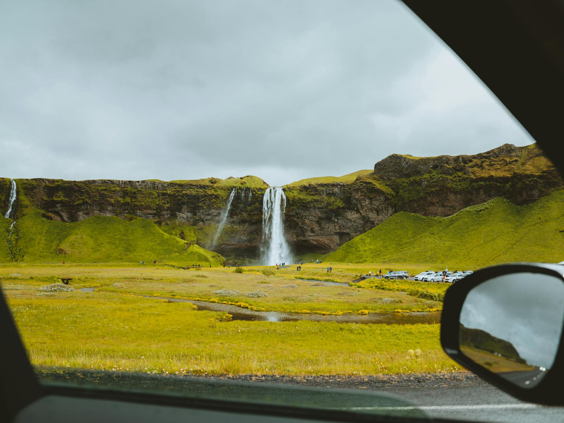 Looking outside the car window, the Seljalandsfoss waterfall on a cliff can be seen in a distance within a green flat landscape 